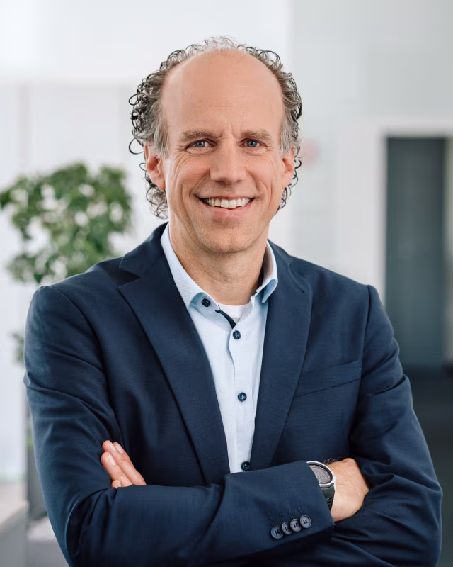 Lächelnder Mann mit Halbglatze, Locken und Anzug steht mit verschränkten Armen in modernem Büro.