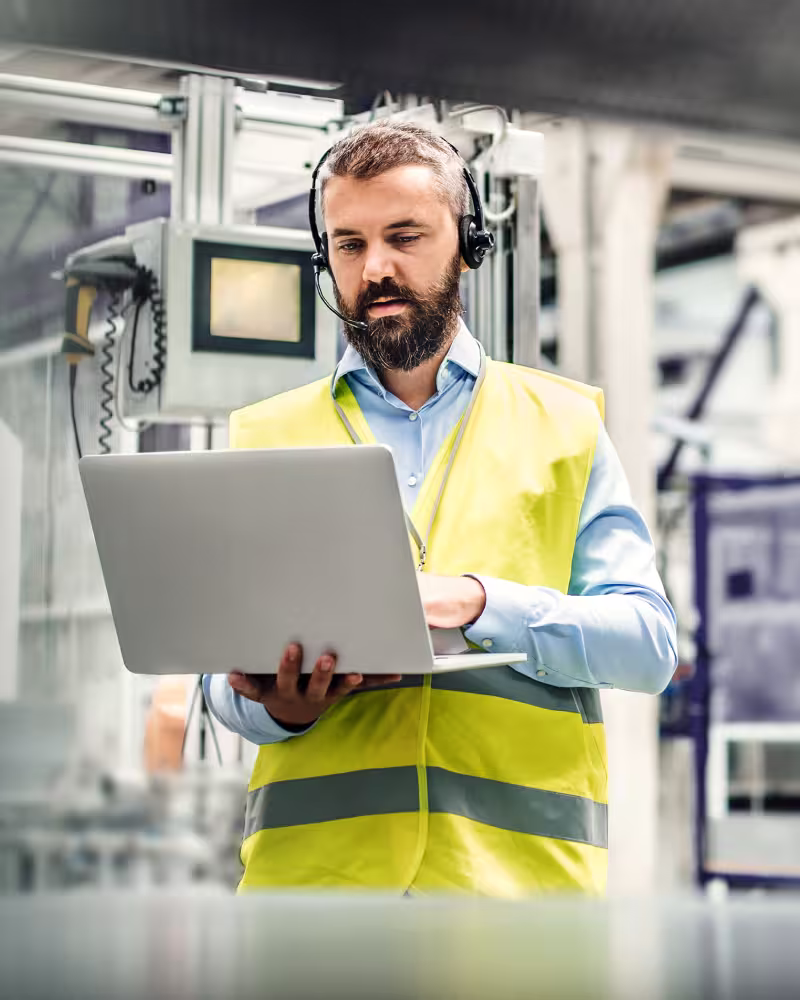 Mann mit gelber Warnweste und Headset bedient Laptop in Industrieumgebung.