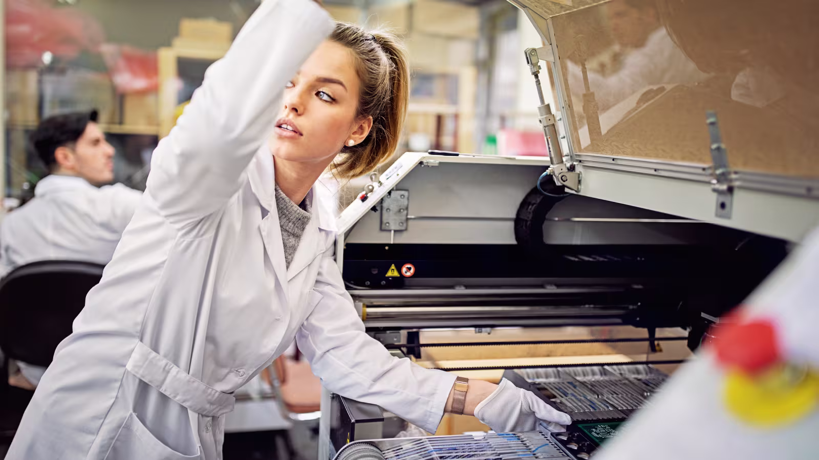 Junge Frau im Labor mit weißem Kittel bedient industrielle Maschine zur Elektronikfertigung.