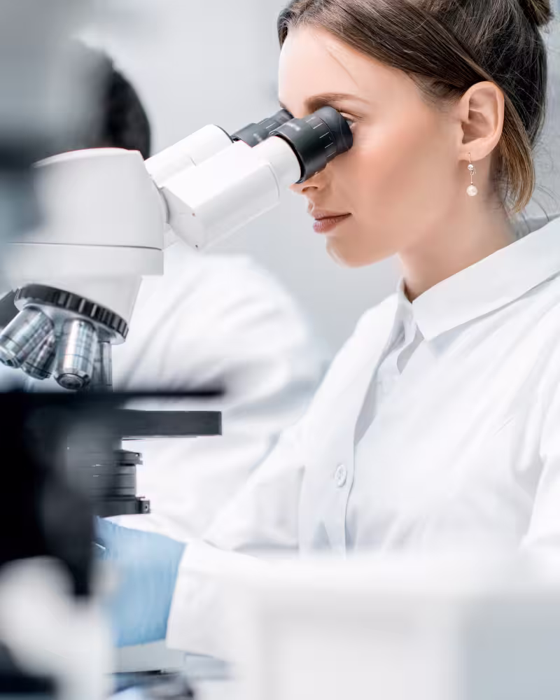 Wissenschaftlerin schaut mit weißem Kittel durch ein Mikroskop im Labor.