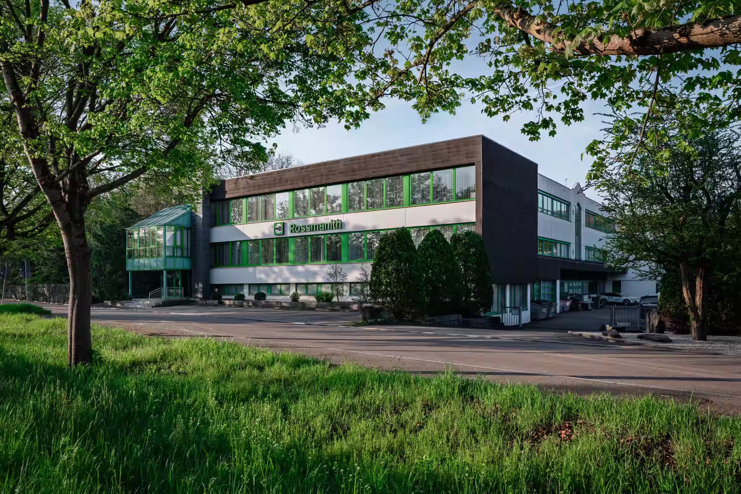 Das moderne Firmengebäude der Firma Rossmanith mit grünen Fenstern und Logo, umgeben von Bäumen und Grasfläche im Vordergrund.